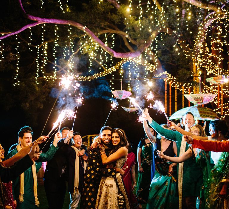 Bride & groom stand outdoors and laugh whilst surrounded by wedding guests who celebrate with sparklers above them