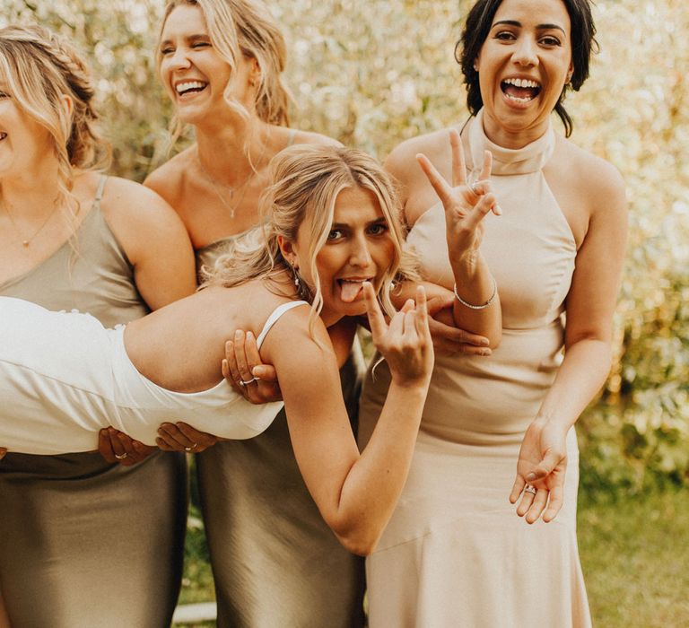 Bridesmaids in sage green and beige bridesmaids dresses hold bride in white Made With Love wedding dress as she sticks out tongue during wedding reception at Anran Devon