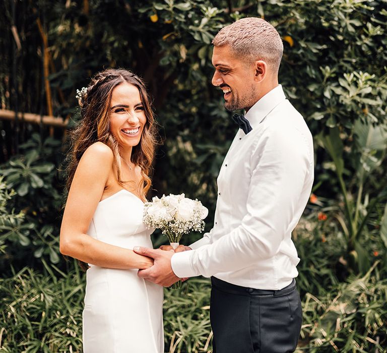 Groom in a white shirt and bow tie holding hands with his bride in a strapless bandeau top and trousers 