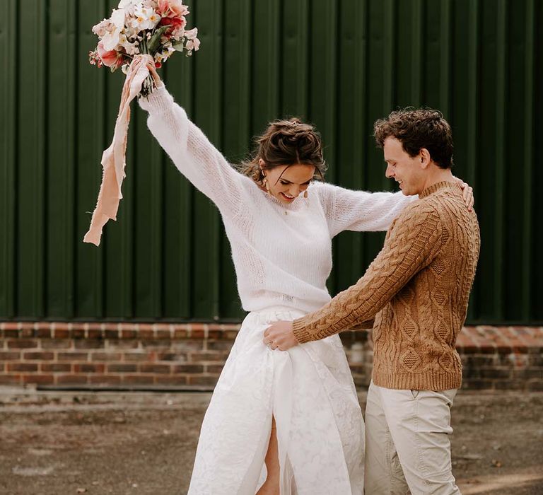 Bride in a lasercut lace wedding skirt, jumper and trainers riding a skate board with her husband in casual wedding attire holding her skirt up 