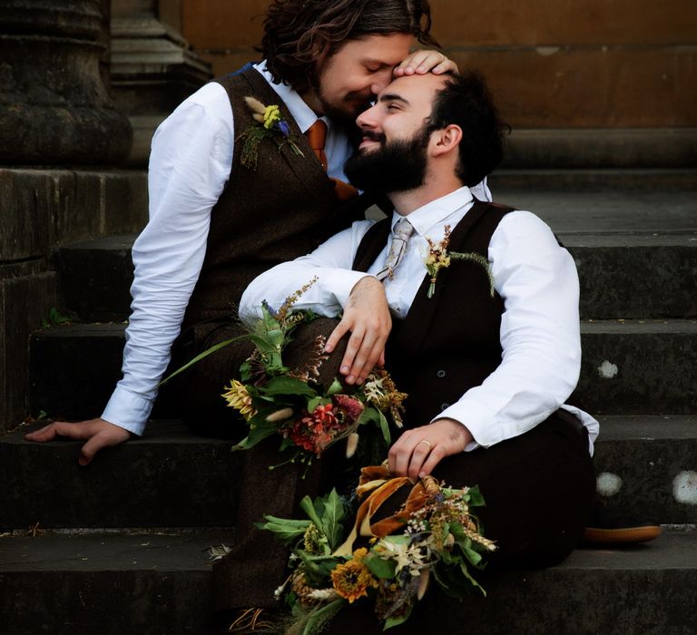 Grooms sit on staircase as they look lovingly at one another