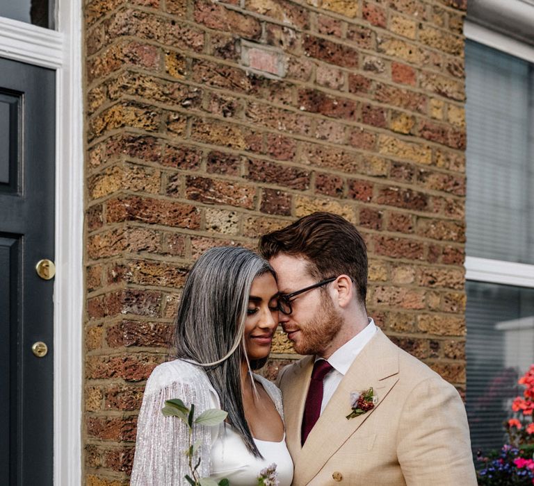 Bride in satin Halfpenny London dress and tasselled bridal cape holding multicoloured bridal bouquet stands close to groom in double breasted linen suit and burgundy tie outside after wedding ceremony at Loft Studios London