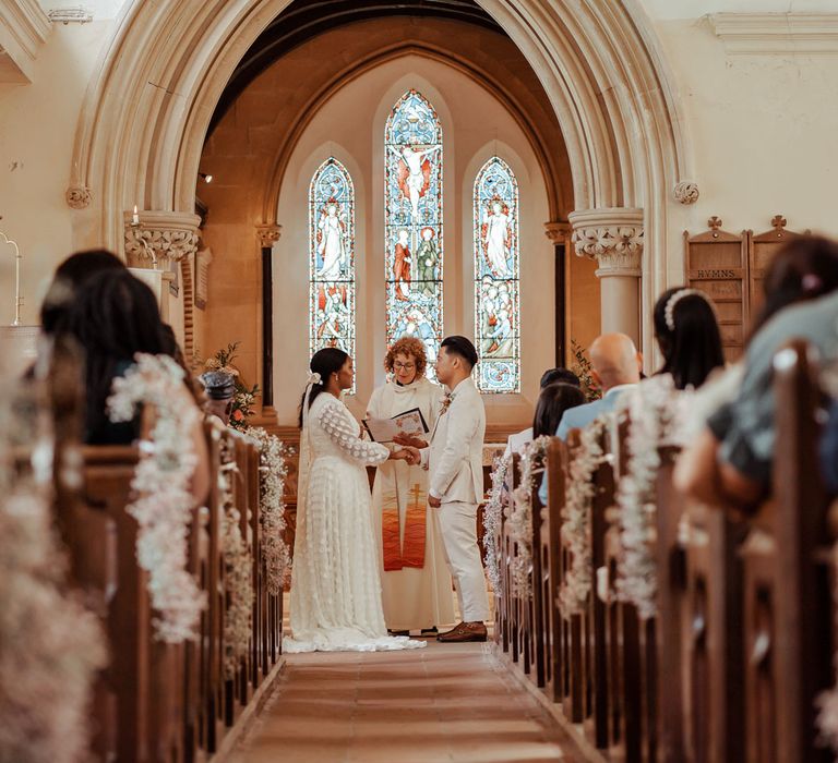 Bride in long mesh sleeve spotted Vagabond wedding dress and bridal hair bow stands facing groom in linen suit at the altar in church during Wasing Park wedding