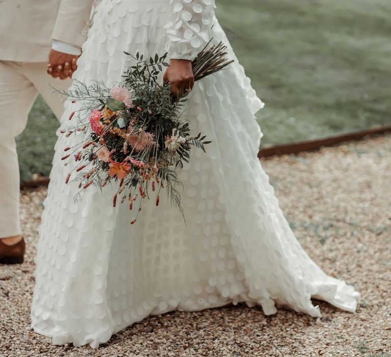 Bride in spotted Vagabond wedding dress holding pink, orange and green mixed bridal bouquet walks holding hands with groom in linen suit and brown shoes at Wasing Park wedding