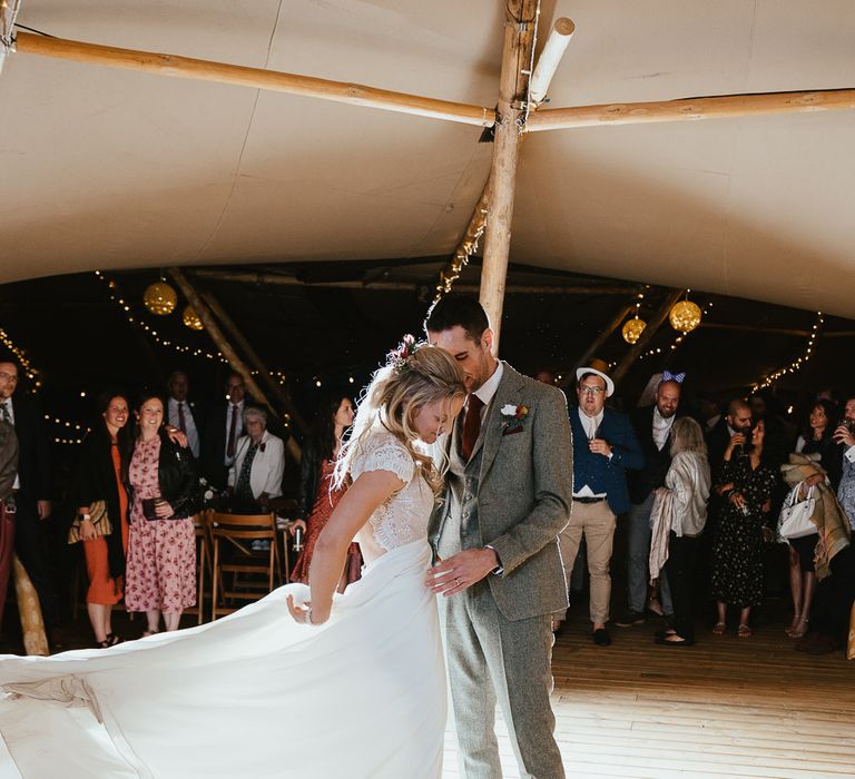 Bride in lace top open back wedding dress flicks the train of her dress as she stands with groom in grey three piece herringbone suit on the dance floor at tipi wedding reception during late summer wedding at Wellington Wood Norfolk 