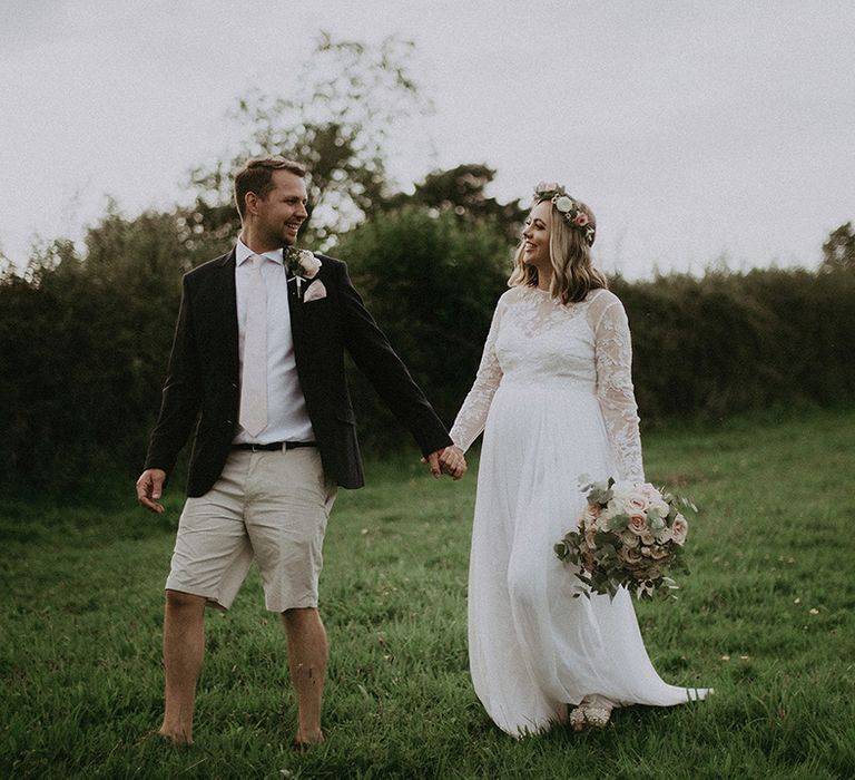 Golden hour bride and groom portrait in a field with groom in beige shorts and navy blue blazer and bride in a lace long sleeve wedding dress and flower crown 