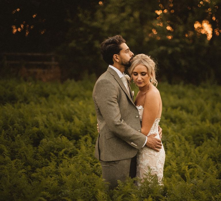 Groom in grey suit hugs bride in off the shoulder Enzoani wedding dress as they stand in field of ferns at Inkersall Grange Farm wedding