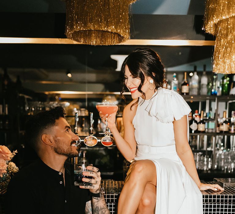 City wedding with groom in a black short sleeve shirt and bride in a stylish wedding dress with boots sitting on the bar drinking cocktails 