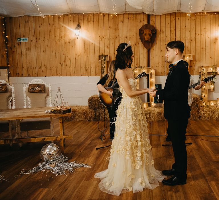 Bride in strapless feathered wedding dress and headpiece holds hands with groom in dark blue suit during rustic wedding ceremony