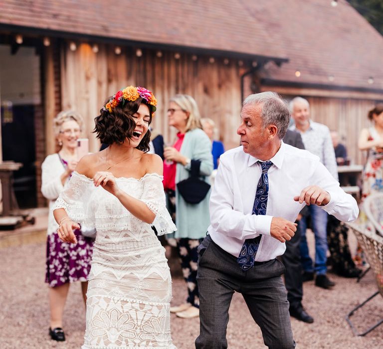 Bride in strapless Grace Loves Lace wedding dress and colourful flower crown dances with guests in grey trousers, white shirt and blue paisley print tie outside at rustic barn wedding