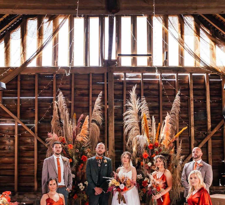 Bride in lace wedding dress stand with groom in green suit with orange bow tie, groomsmen in grey suits and orange ties and bridesmaids in orange satin bridesmaid dresses