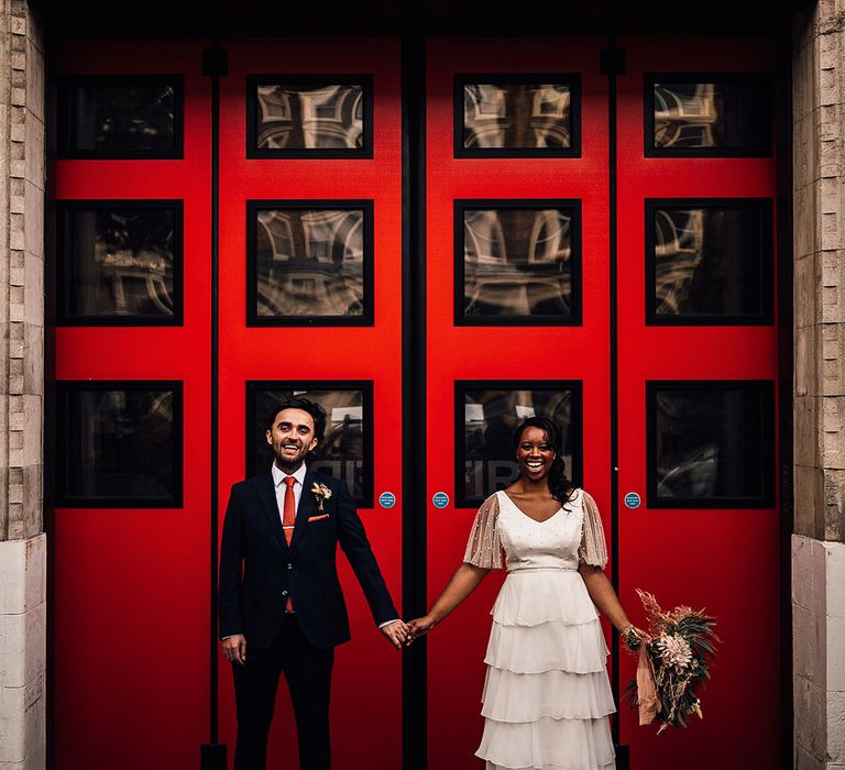 Black bride in a layered skirt and pearl top holding hands with her groom in a navy suit in front of Brixton station's red doors 