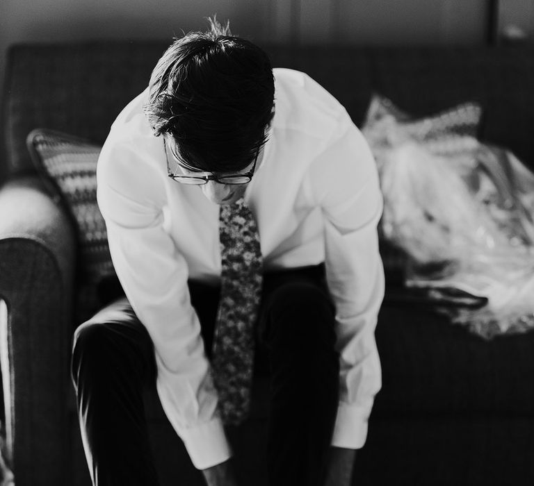 Groom in white shirt and floral tie puts on shoes before his wedding