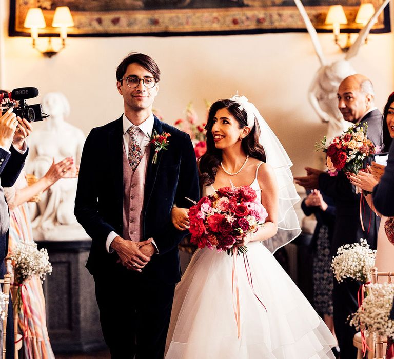 Bride in A line tulle skirt wedding dress and veil holding mixed red, white and pink bridal bouquet looks at groom in dark suit, baby pink waistcoat and floral tie as they walk down the aisle together