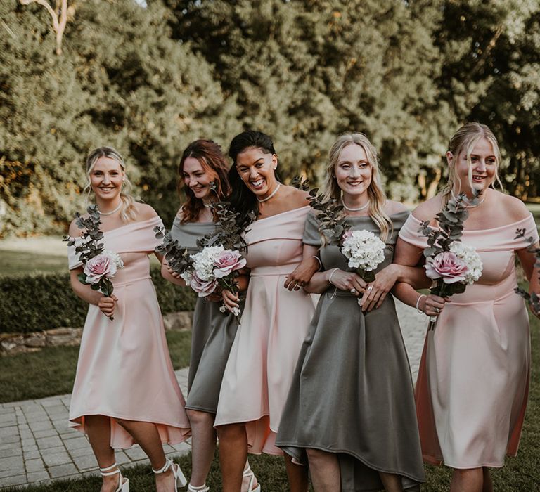 Bridesmaids wear off the shoulder tea length bridesmaids gowns in sage green and pastel pink whilst holding floral bouquets