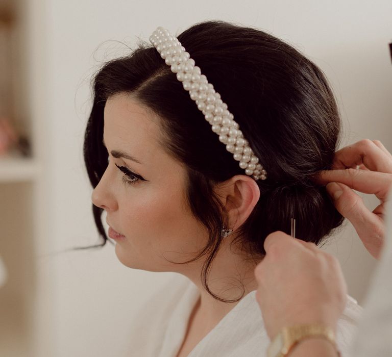 Bride gets her hair done into a sleek updo decorated with a trendy pearl headband accessory 