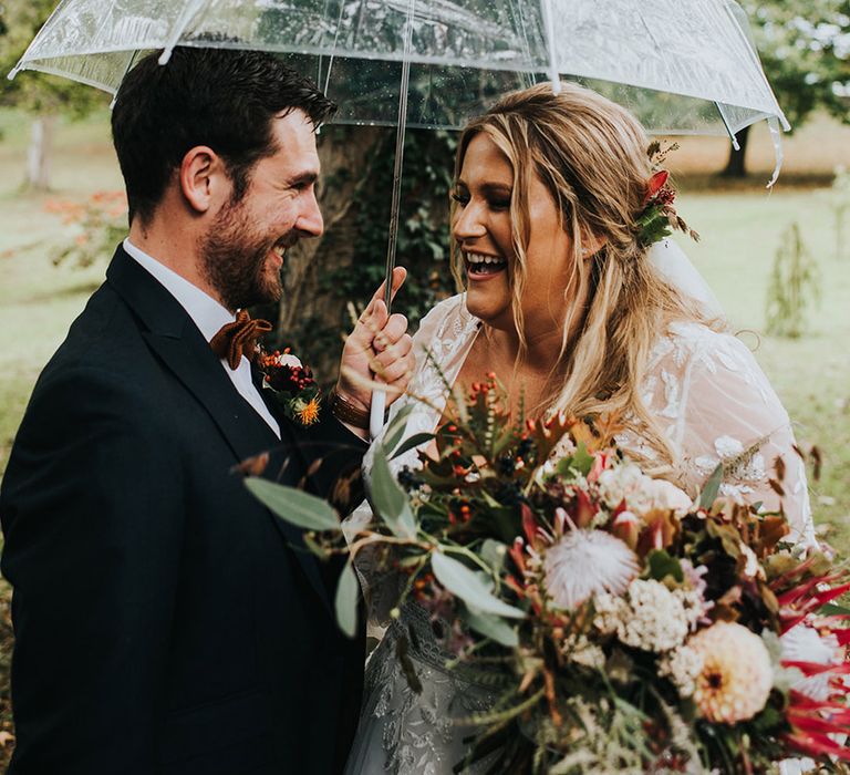 Rainy wedding day with bride and groom standing under a clear umbrella as the bride holds her autumnal bouquet with proteas 