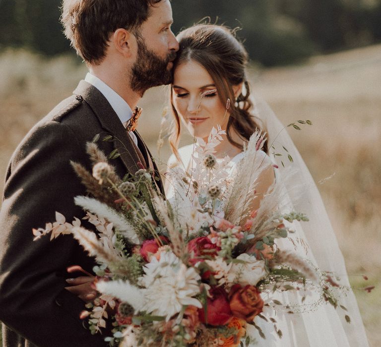 Groom in paisley bow tie kisses bride on the head with large autumnal orange and white bouquet with pampas grass 