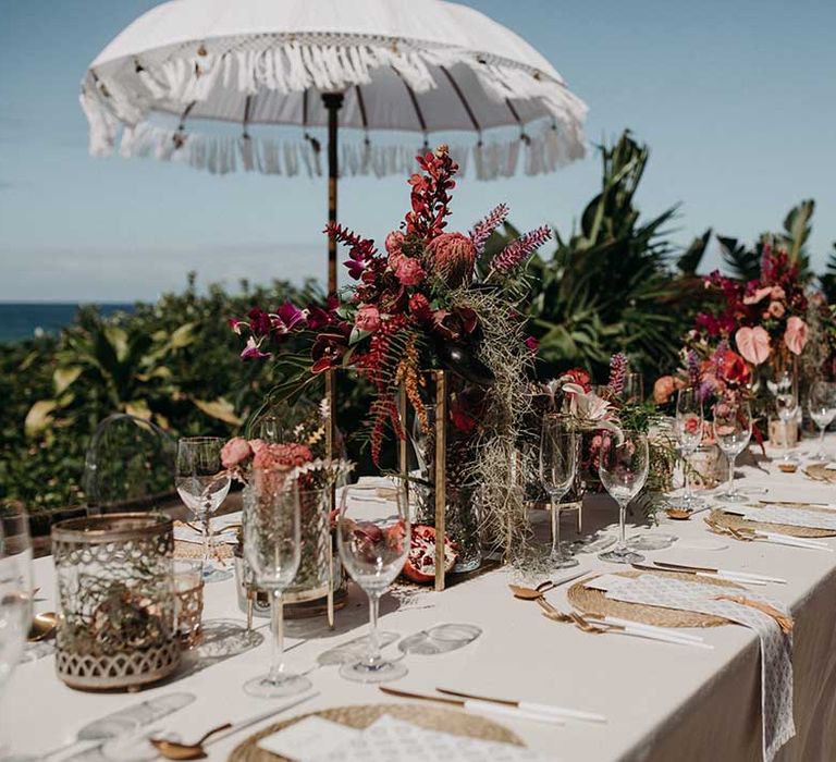 Outdoor table setting for destination wedding with woven placemats, red and pink flowers and white umbrella with tassels 