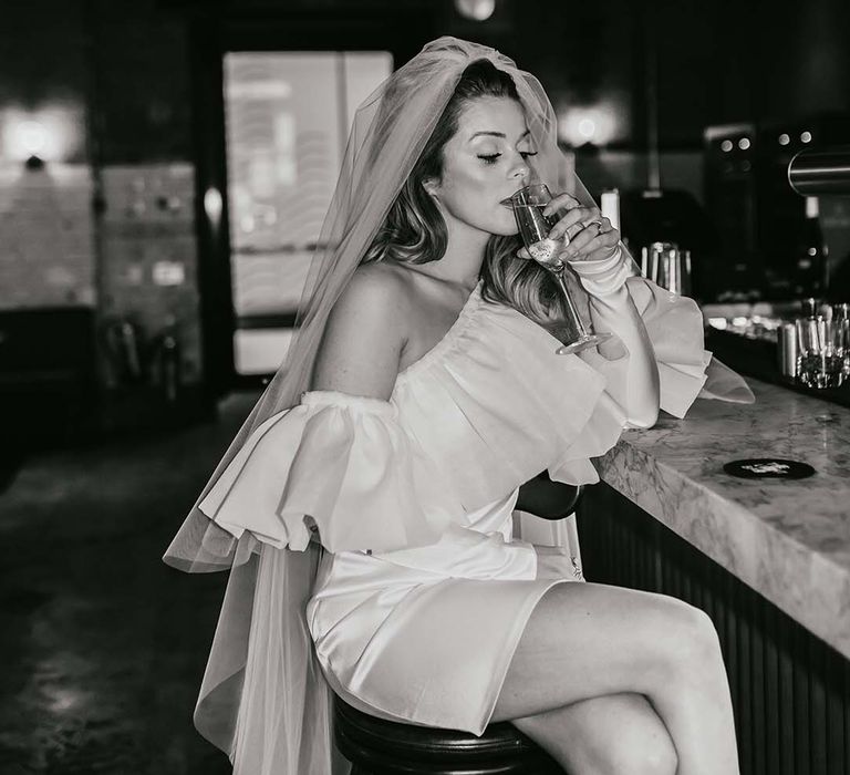 Bride in a ruffle off the shoulder short wedding dress sitting at the bar drinking Champagne 