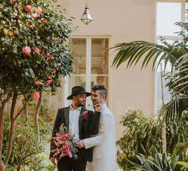 Groom in a black suit and fedora hat standing with his partner in a white groom suit and cape holding a vibrant red bouquet 