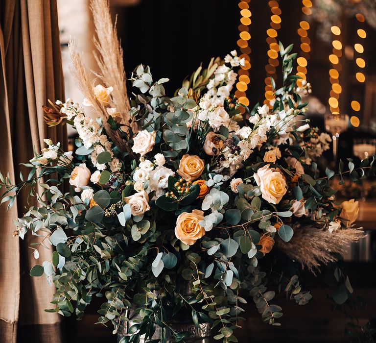 Large white and orange flower arrangement with dried grass in vintage milk churns for rustic decor at barn wedding 