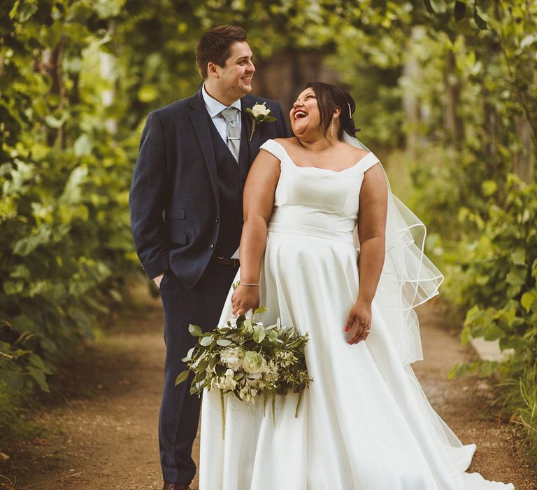 Bride in bardot wedding dress with dangly earrings laughs with groom in three piece blue suit 