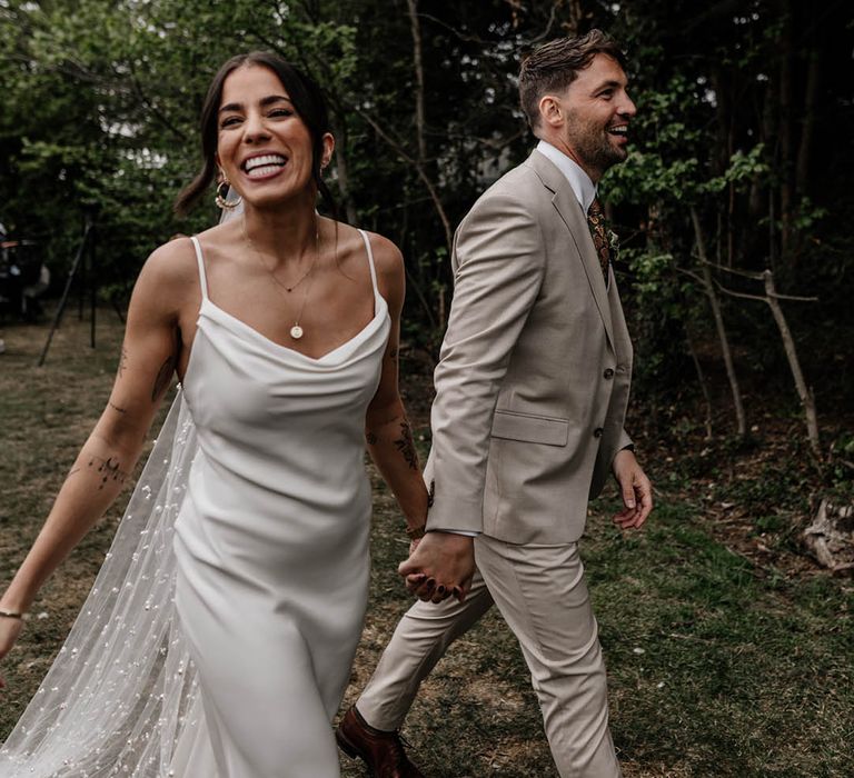 Groom in a stone coloured suit holding hands with his bride in a satin slip wedding dress and pearl veil 