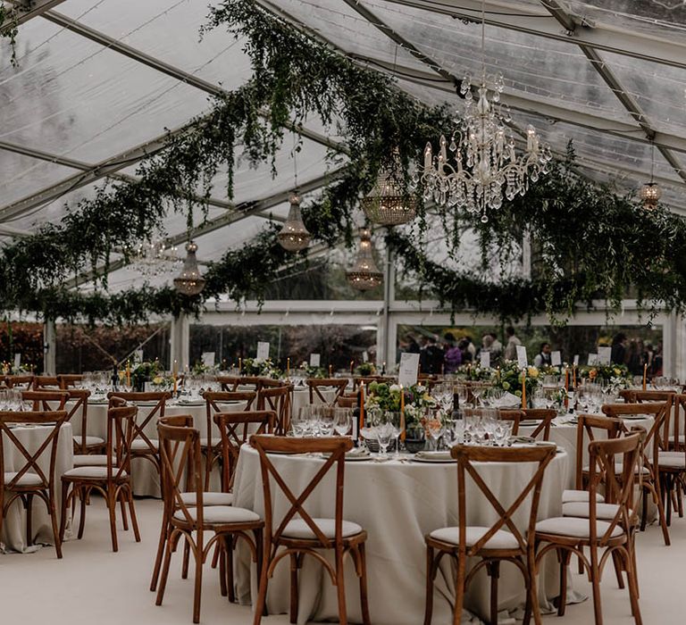 Clear glass marquee wedding reception with wooden furniture, greenery decor and handing chandelier 