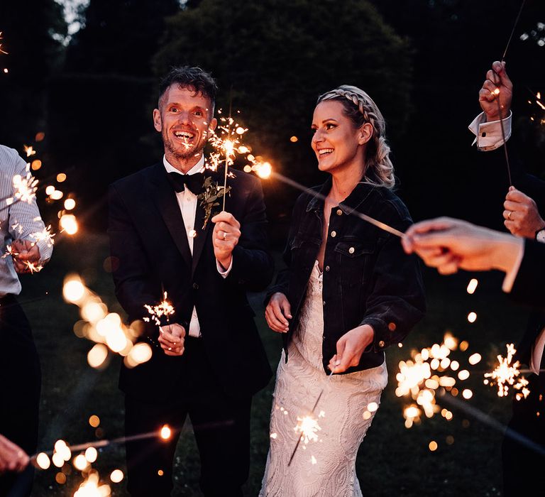 Bride and groom wave sparklers with their guests with the bride wearing a black denim cover up 