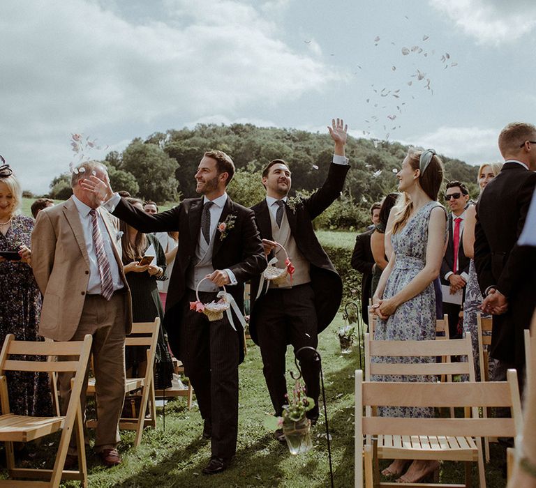 Groomsmen in morning suits walk down the aisle with wicker baskets with pink ribbons throwing out petals 