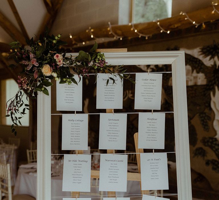 Large white photo frame seating chart with white cards attached with string and mini pegs on a wooden easel 