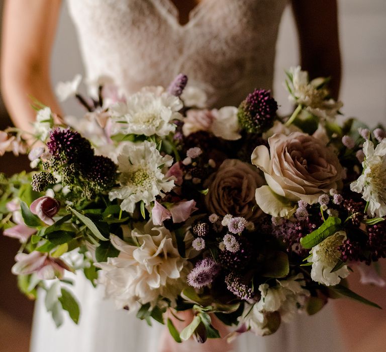 Bride wears lace wedding gown with N-neck shape to front and holds purple bridal bouquet 