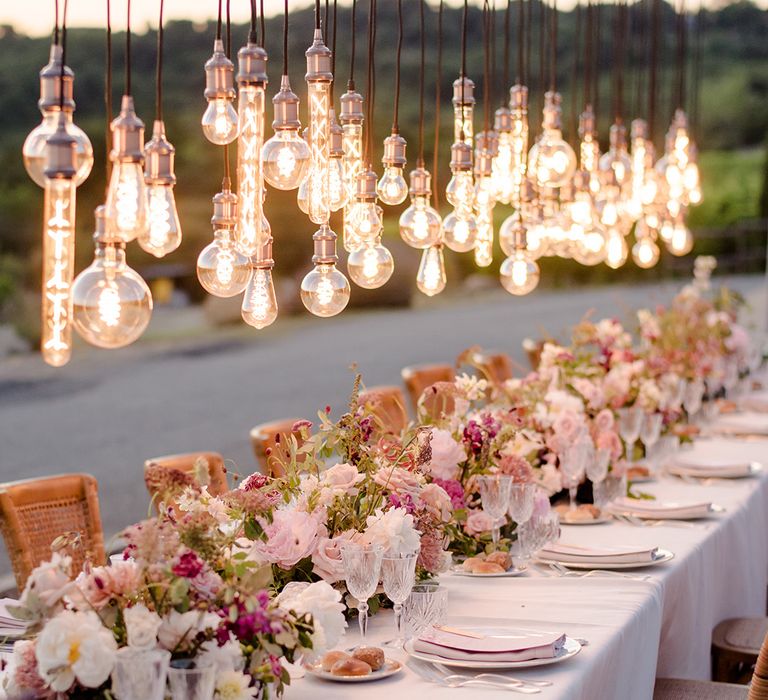 Edison bulbs hang above head table during outdoor wedding reception in Tuscany complete with pale pink and purple floral bouquets 