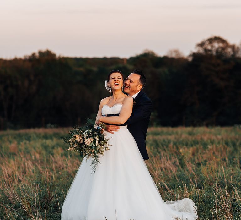 Groom embraces the bride from behind as they both laugh together during sunset on their wedding day for couple portraits