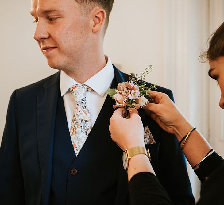 Groom wears three piece suit complete with colourful tie as wedding guest places floral buttonhole in suit jacket 