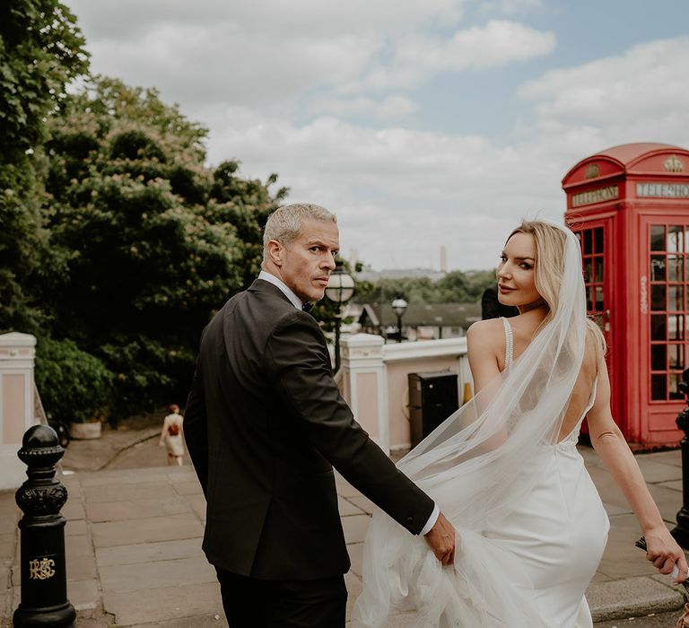 Bride and groom walk and look back at the camera with groom in black tie and bride wearing a long veil 