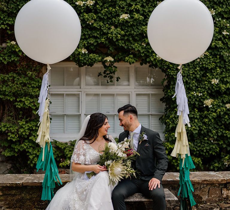 Bride & groom sit on bench outdoors complete with large white balloons and different coloured tassels 