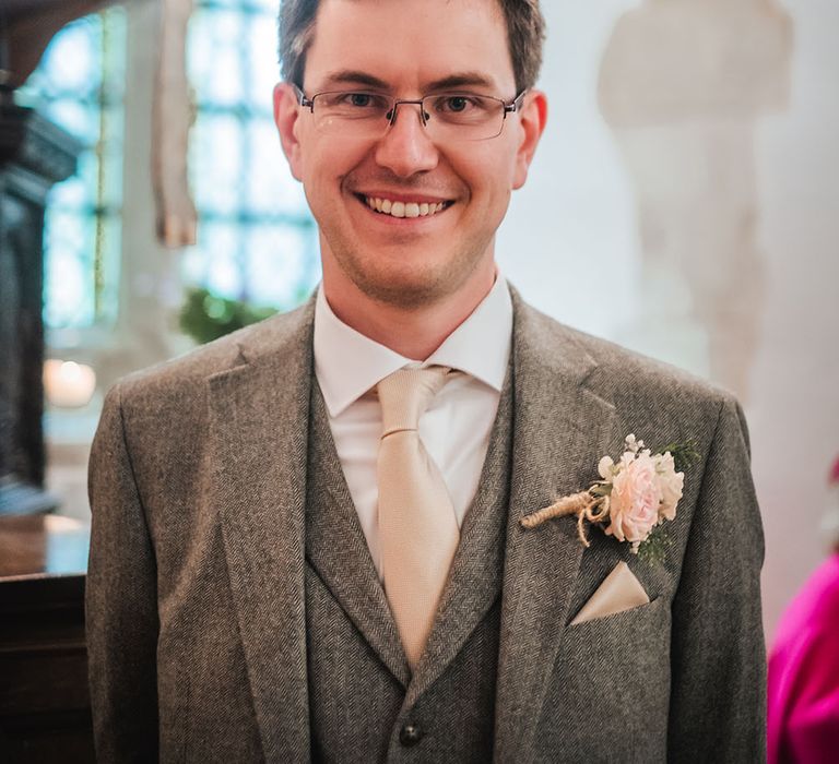 Groom wears grey three piece suit with pale pink tie and floral buttonhole tied with brown string