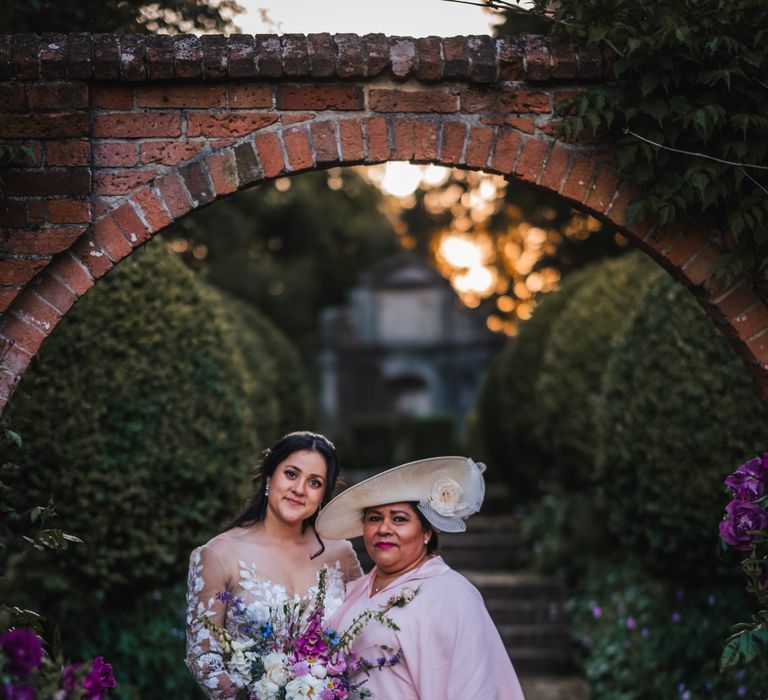 Bride stands outdoors with her mother who wearing pastel pink dress and white hate finished with roses 