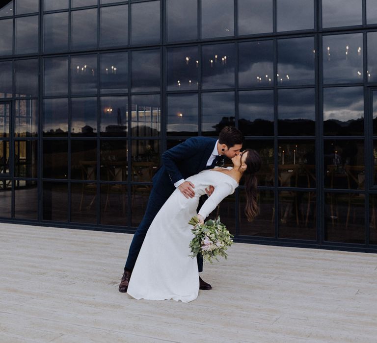 Groom in blue suit leans the bride down for a kiss in front of floor to ceiling windows 