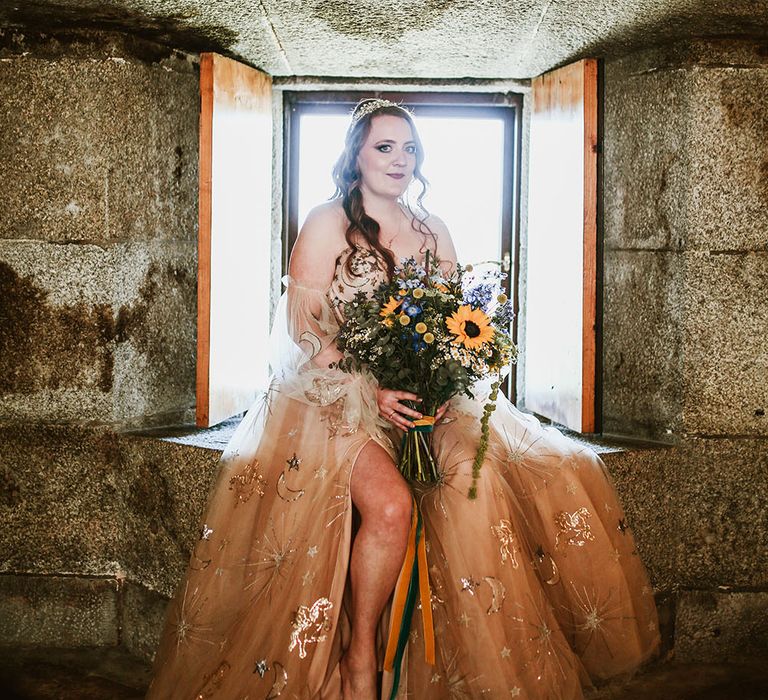 Bride wears pink starry wedding dress with off-the-shoulder sheer puffed sleeves and holds oversized floral bouquet with sunflowers and tied with green and orange velvet ribbon