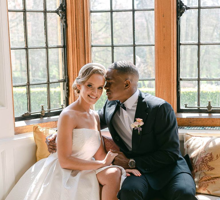 Groom sits on the window seat with the bride and rests his forehead against her as she smiles at the camera 