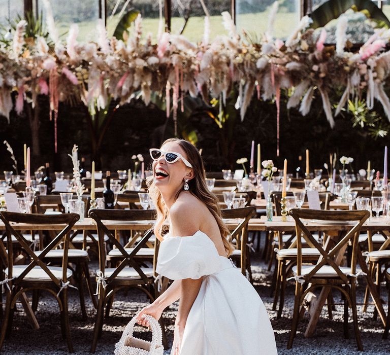 Bride in a puff sleeve wedding dress holding a pearl handbag with white sunglasses smiling at her pastel theme wedding breakfast