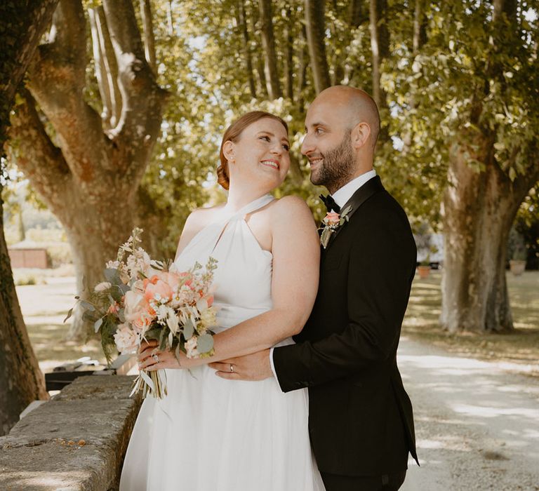 Bride holds peony floral bouquet and looks back lovingly at her groom after outdoor wedding ceremony in France with classic styling