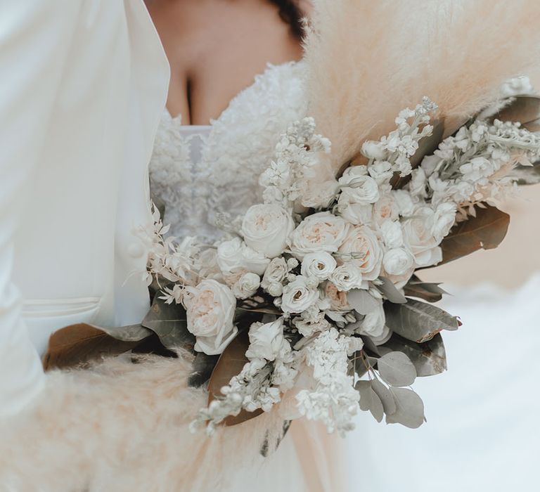 Bride holds oversized pampas grass bouquet as she stands at the sea front with her groom on their wedding day