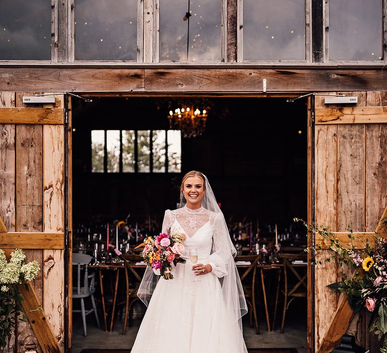 Bride in a long sleeve delicate lace wedding dress stands at the entrance to the reception holding a glass of champagne with colourful wedding flowers