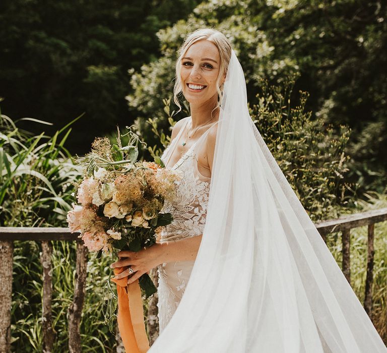 Bride with a long veil in a lace wedding dress wearing a blue pendant necklace holding an orange coloured bouquet 