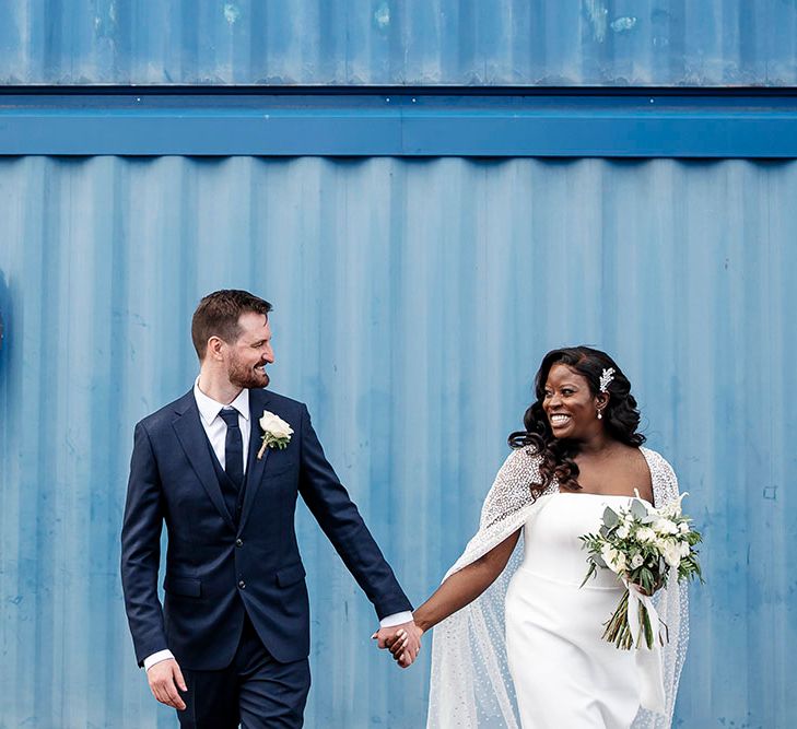 Bride & groom hold hands and look lovingly at one another in front of light blue wall 