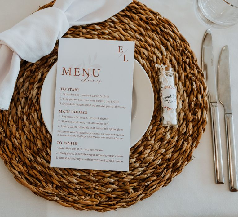 Minimalist white wedding dinner menu on wicker place mat with small wedding favour, tube of mints, to the right